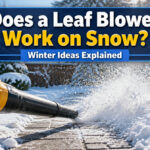 A leaf blower positioned on a snowy surface with a garden shed in the background.
