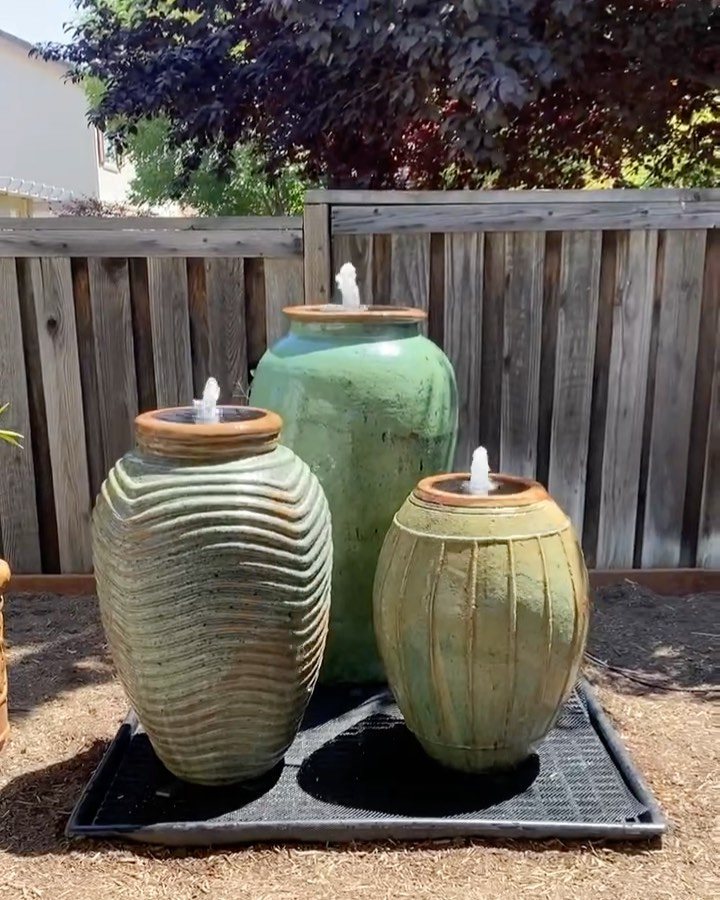 Three green ceramic pot fountains bubbling water on a black tray in a backyard.